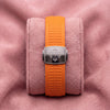 Orange textured Aquanaut strap with silver-toned cross-emblem clasp on a pink cushion