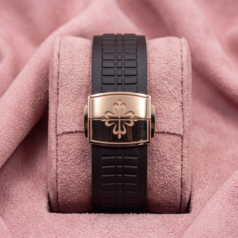 Dark textured Aquanaut strap with rose-toned cross-emblem clasp on a pink cushion